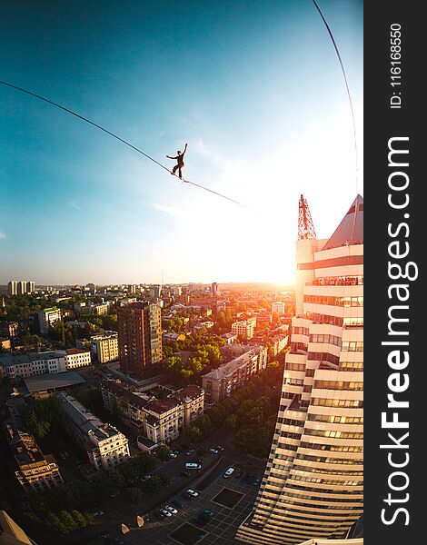 A man walks along a line stretched between two buildings at sunset. A man catches balance at altitude. Highline over the city.