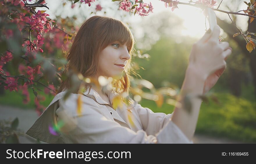 Young attractive red-haired woman taking photos of spring flowers of cherry or sakura blossoms on smartphone at sunset in park in Odessa