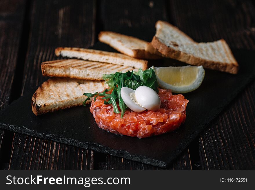 Close-up of salmon tartare served with quail egg, butter, bread and lemon on wood background