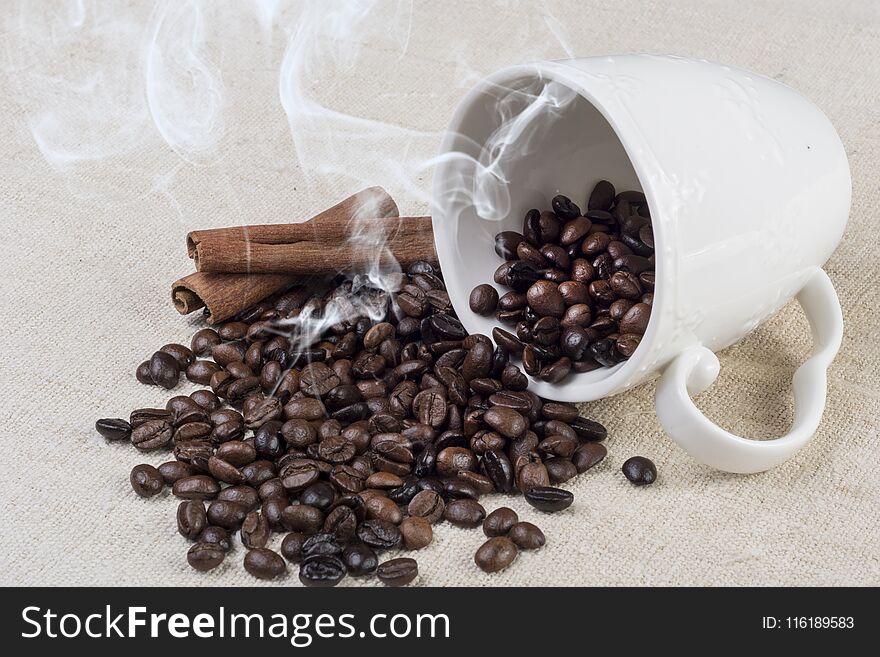 Black Fresh roasted coffee bean smoke and spilled from white coffee cup and cinnamon on retro fabric tablecloth on desk. Close up, selective focus. Retro food and drink concept image. Black Fresh roasted coffee bean smoke and spilled from white coffee cup and cinnamon on retro fabric tablecloth on desk. Close up, selective focus. Retro food and drink concept image.