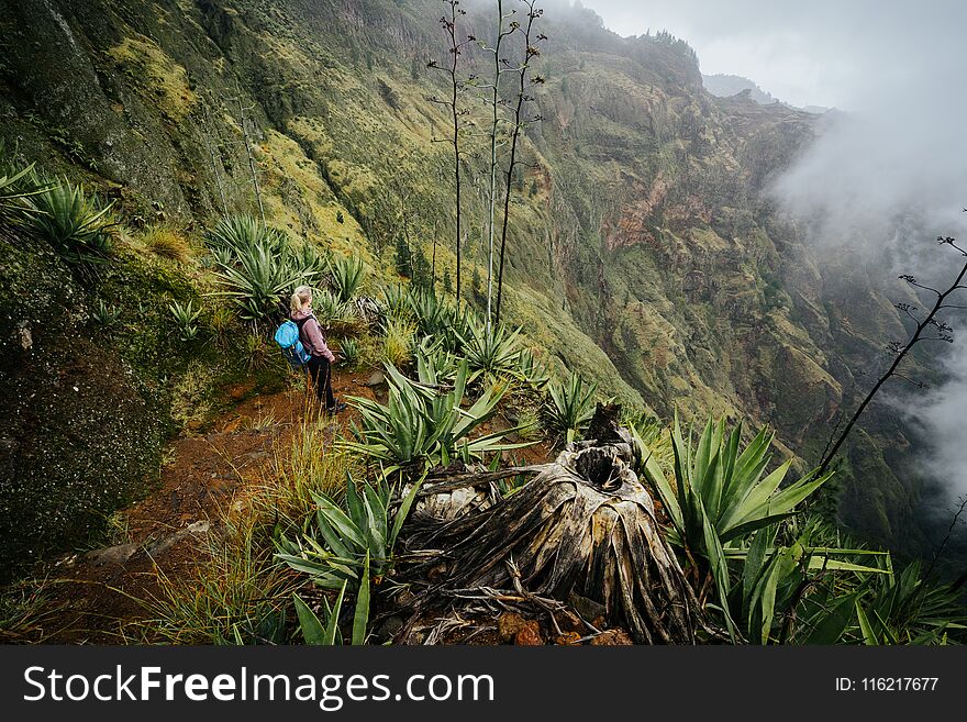 Female traveler staying on the cove volcano edge above the foggy green valley overgrown with agaves Santo Antao island