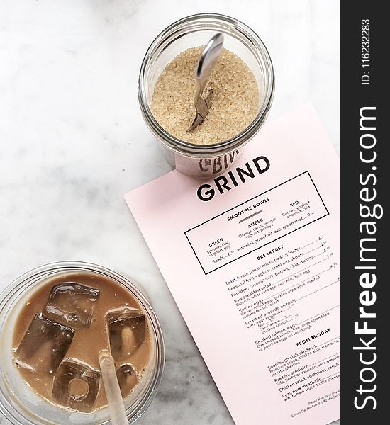 Clear Drinking Glass Filled With Tea and Ice Cubes With Clear Plastic Straw Beside Clear Glass Canister Filled With Brown Sugar