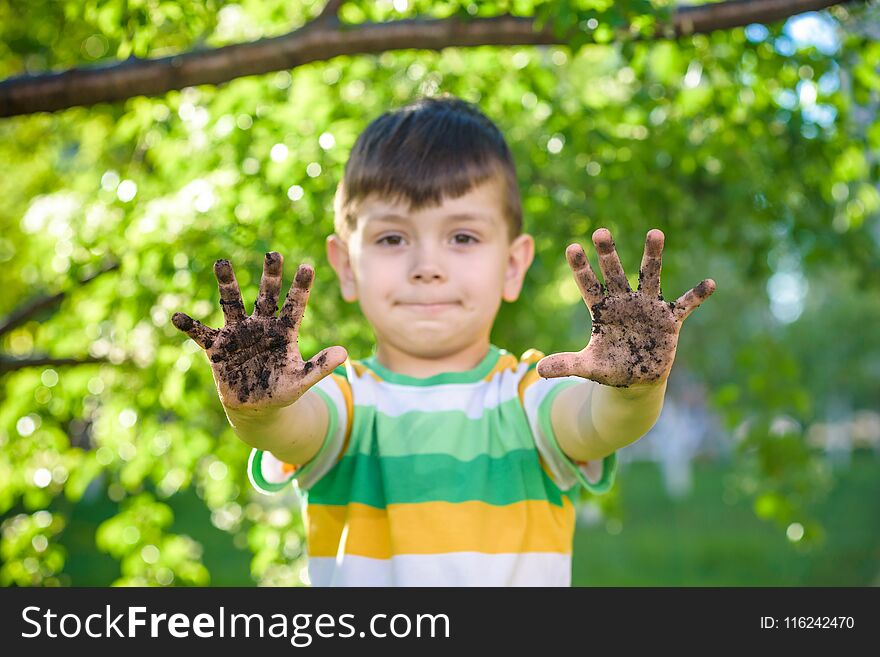 A young Caucasian boy showing off his dirty hands after playing in dirt and sand outdoors sunny spring or summer evening on blossom trees background. happy childhood friendship concept.