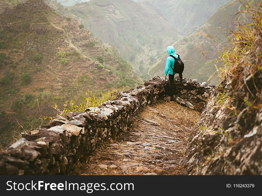 Hiker with backpack on the mountain edge cobbled path looking down the valley. Rocky terrain of high mountain ranges and