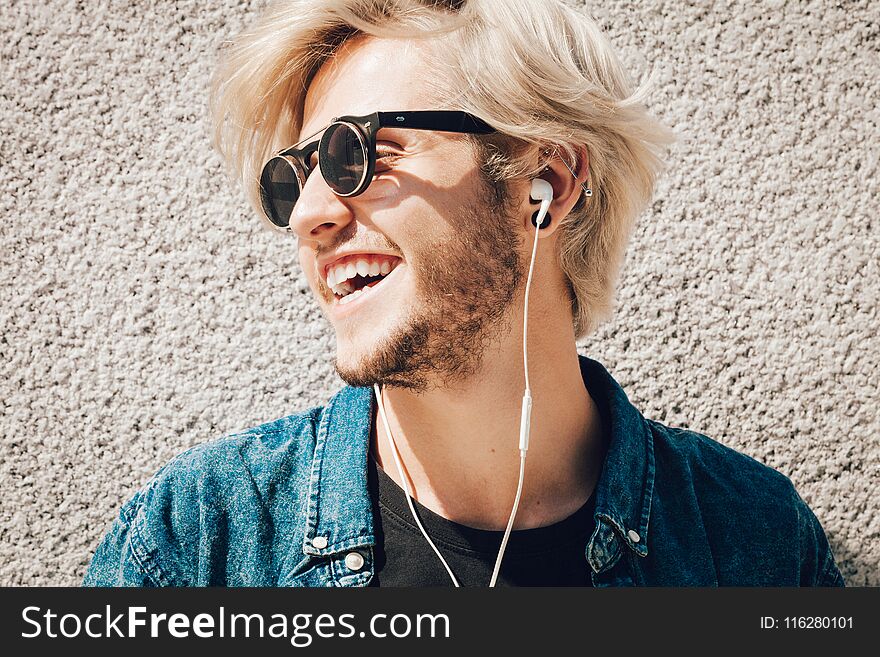 Men fashion, technology, urban style clothing concept. Hipster smiling guy standing on city street wearing jeans outfit listening to music through earphones. Men fashion, technology, urban style clothing concept. Hipster smiling guy standing on city street wearing jeans outfit listening to music through earphones
