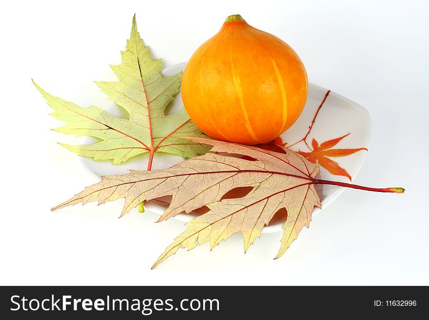 The small orange pumpkin is on a plate near to maple leaves. The small orange pumpkin is on a plate near to maple leaves