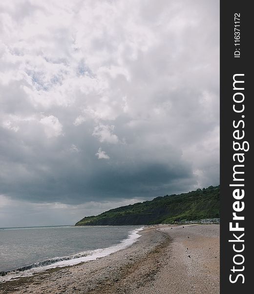 Beach Sand Near Body of Water and Green Mountain Under Cloudy Sky