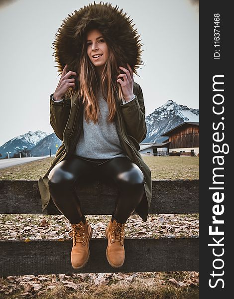 Woman Wearing Brown and Green Parka Sitting on Gray Wooden Fence at Daytime