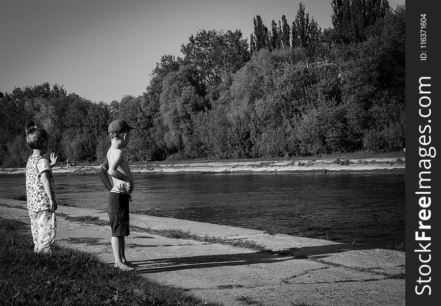 Grayscale Photo Of Oy Boy And Girl Standing Near Body Of Water