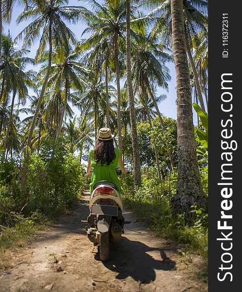 Back view of young happy tourist woman with hat riding scooter motorbike in tropical paradise jungle with blue sky and palm trees exploring trip destination and Summer holidays travel in Asia