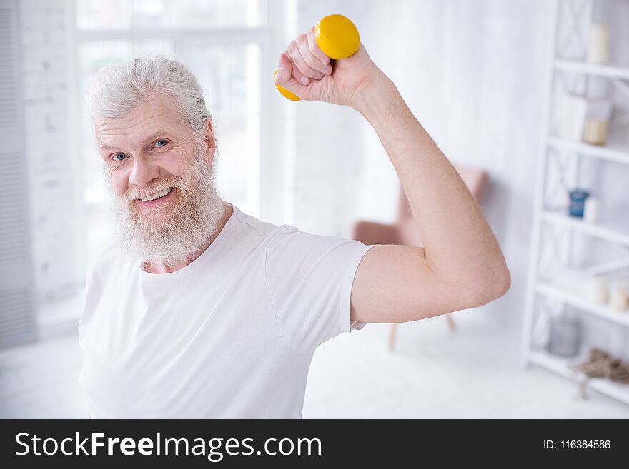 Pleasant exercise. Cheerful elderly man smiling at the camera and raising hand while holding a dumbbell. Pleasant exercise. Cheerful elderly man smiling at the camera and raising hand while holding a dumbbell
