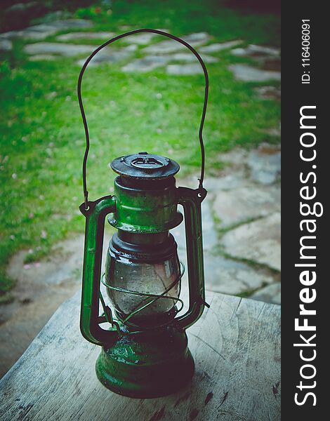 Photo depicts metal rusty lantern. An old oil kerosene green lampplaced on a wooden table in the garden. Summertime, footpath paved with stone and blurred nature on the background.