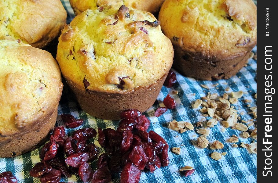 Freshly baked cranberry oatmeal muffins closeup with loosely scattered oats and dried cranberries on a blue-green gingham cloth background. Freshly baked cranberry oatmeal muffins closeup with loosely scattered oats and dried cranberries on a blue-green gingham cloth background