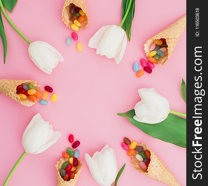 Frame made of sugar candy in waffle cones and white tulip flowers on pink background. Flat lay, top view