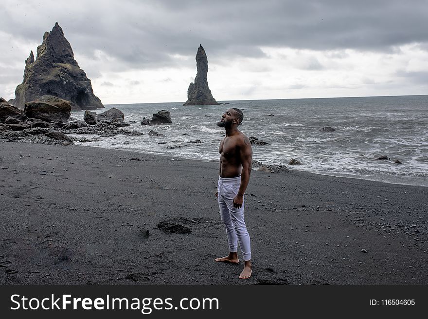 Man in White Jeans Stands on Grey Sand in Beach Under Grey Clouds
