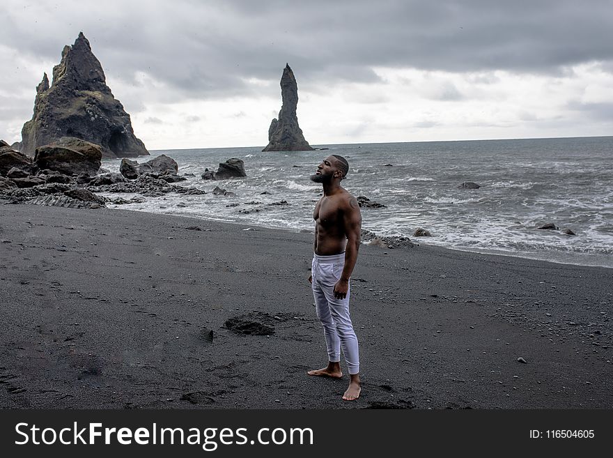 Man In White Jeans Stands On Grey Sand In Beach Under Grey Clouds