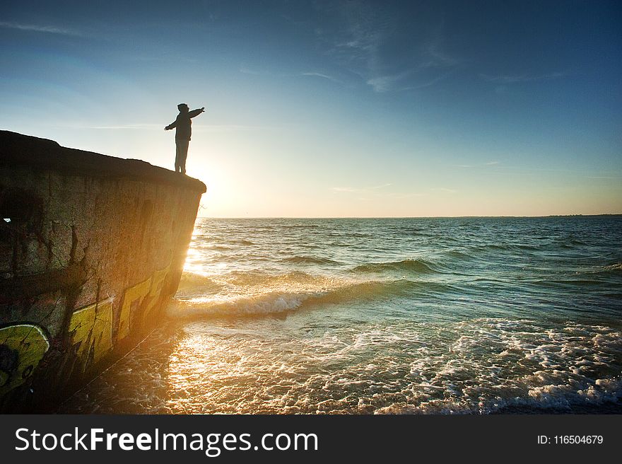 Silhouette of Person on Cliff Beside Body of Water during Golden Hour