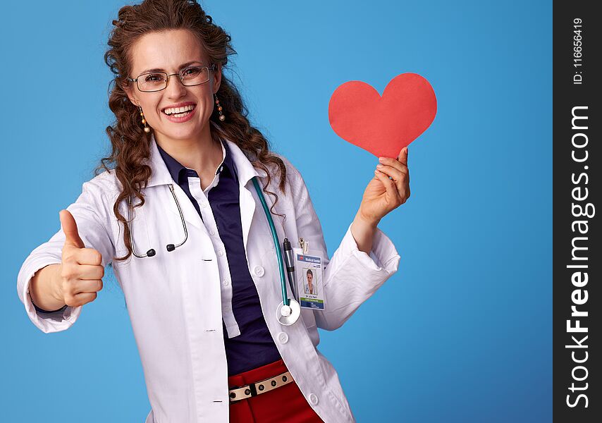 Smiling medical practitioner woman in white medical robe showing thumbs up and heart isolated on blue background. Smiling medical practitioner woman in white medical robe showing thumbs up and heart isolated on blue background