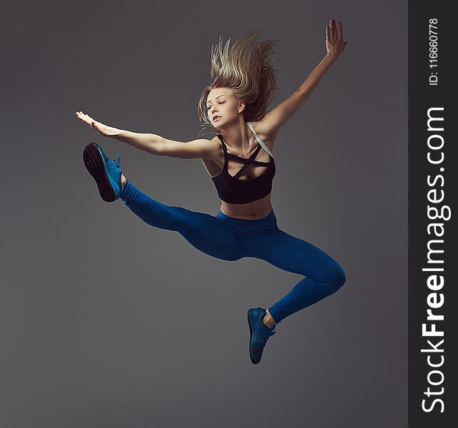 Young blonde ballerina in sportswear dances and jumps in a studio. Isolated on a gray background.
