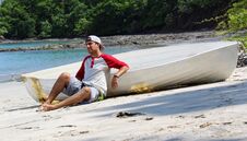 Good Looking Castaway Man Sitting In The Beach By A Wrecked Boat Waiting For Help With Ocean And Jungle In The Background. Royalty Free Stock Image