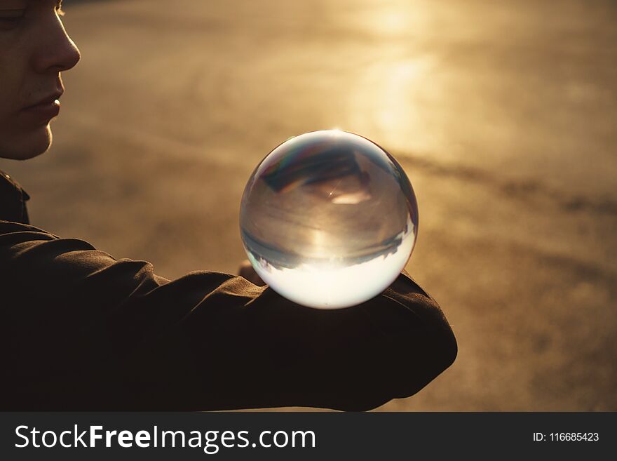 Contact juggling. Guy balances with glass ball on bent elbow. Inverted panorama of city in reflection of ball. Mastery of representation. Close-up photo at sunset.