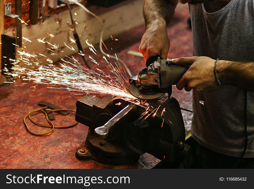 Blacksmith polishes crossguard of sword. Man is working in workshop. He holds a grinder in his hands.