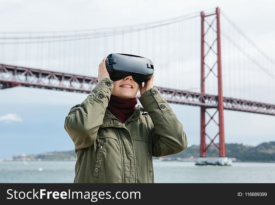 Young beautiful girl wearing virtual reality glasses. 25th of April bridge in Lisbon in the background. The concept of modern technologies and their use in everyday life.