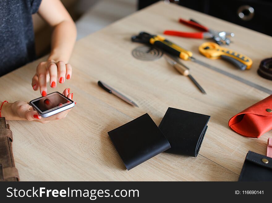 Female hands with red nails at leather atelier using smartphone, handmade wallets and tools on table. Concept of handicraft business and craftswoman with modern technology.