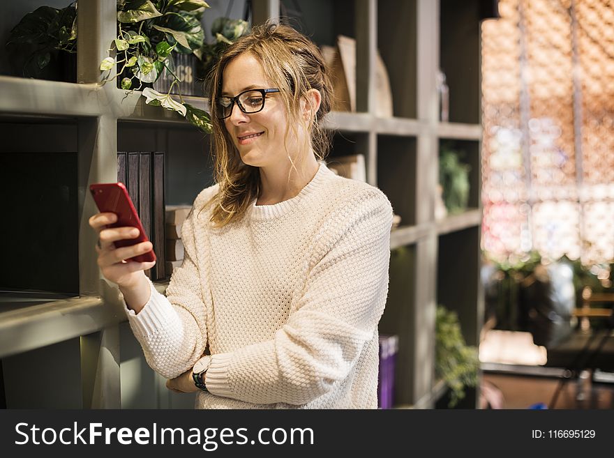 Selective Focus Photography of Woman Using Smartphone Beside Bookshelf