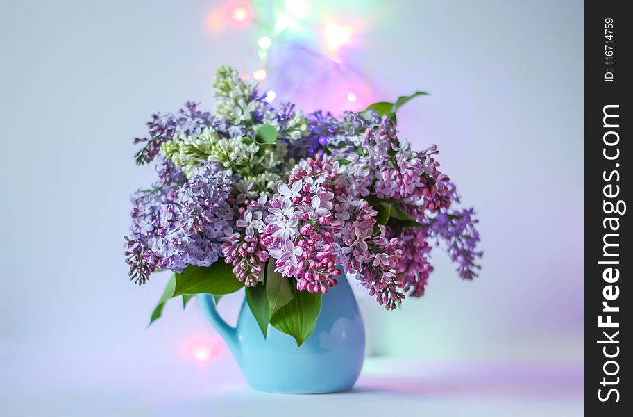 Beautiful bouquet of fragrant purple flowers in blue ceramics vase on light background with festive garland colorful lights. Syringa vulgaris or lilacs plant. Beautiful bouquet of fragrant purple flowers in blue ceramics vase on light background with festive garland colorful lights. Syringa vulgaris or lilacs plant.