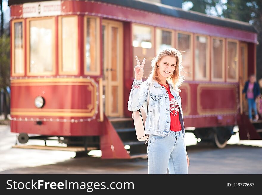 Beautiful girl in a blue denim jacket and jeans against a red tram. Woman shows the world with gestures in the sunset sun with a backpack on the bus background