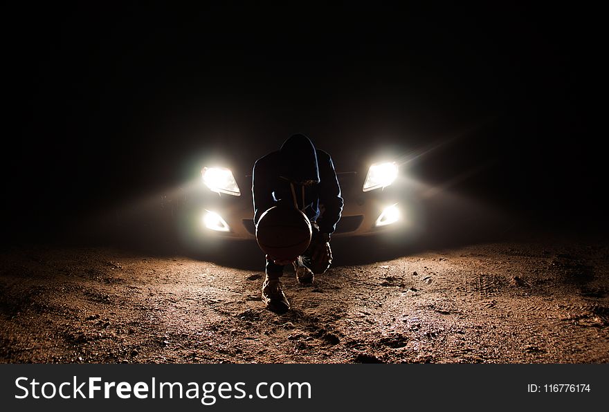 Man Against the Vehicle Headlight Squatting Holding Ball