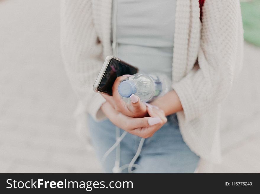 Selective Focus Photography of Woman in White Cardigan Holding Water Bottle and Black Smartphone