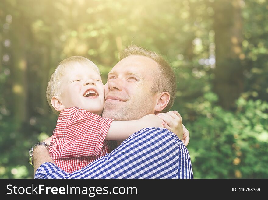 Father and son hugging, smiling and looking at the sun and screwing up one`s eyes in the garden. Masculinity concept. Blondy people. Family. Father and son hugging, smiling and looking at the sun and screwing up one`s eyes in the garden. Masculinity concept. Blondy people. Family.