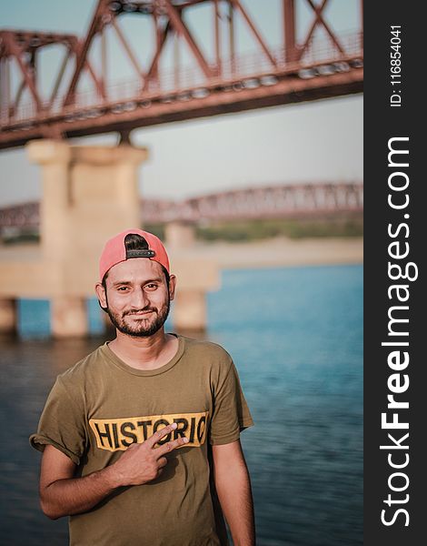 Man Wearing Gray Historic-printed T-shirt and Red Snapback Cap Taking Photo Beside Body of Water