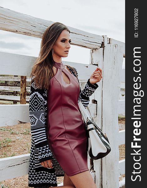 Woman Wearing Maroon Leather Dress And Cardigan Leaning On White Plank Wall