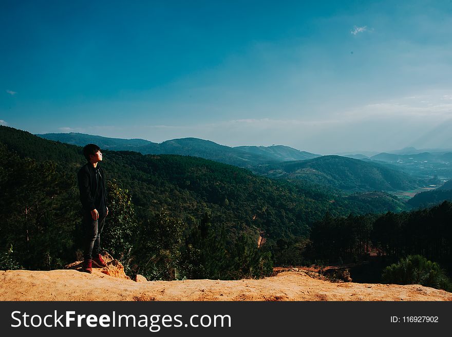 Man Stands on Cliff With Green Mountains on Background