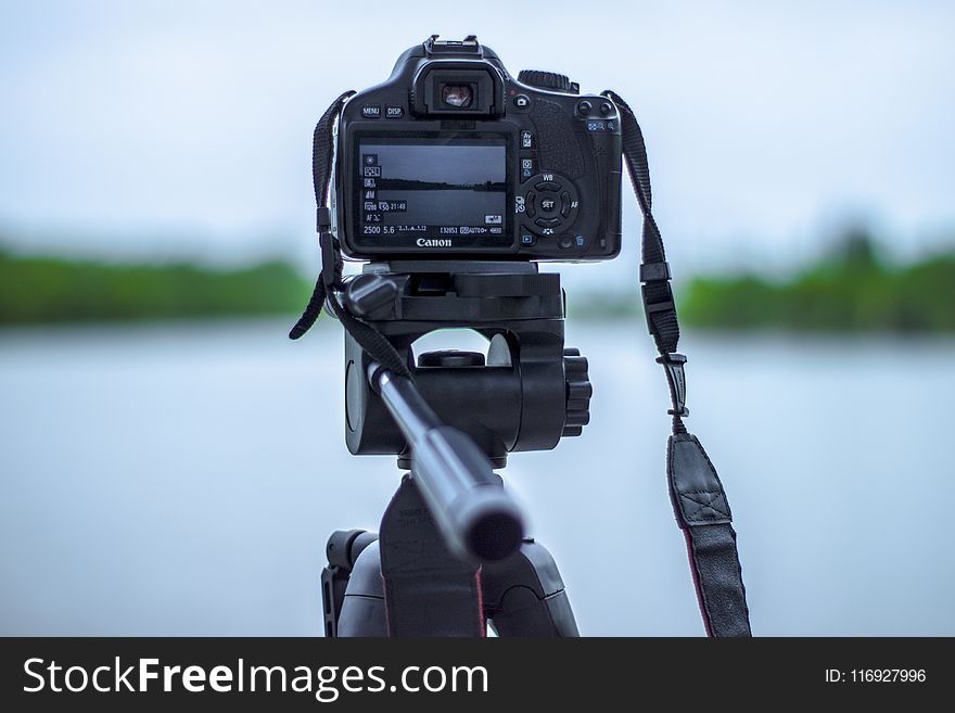 Selective Focus Photo Of Black Canon Camera On Tripod Stand In Front Of Body Of Water Photo