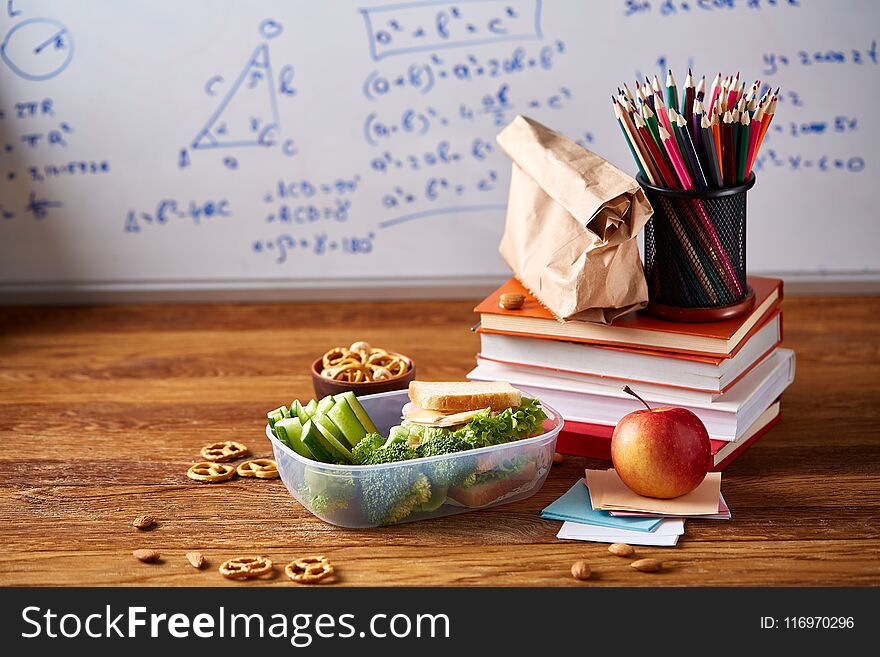 Back to School concept, school supplies, assortment of biscuits, paper paper with food and transparent lunchbox full of sandwich, fruits and vegetables togather with assortment of biscuits over white chalkboard, selective focus, close-up. Back to School concept, school supplies, assortment of biscuits, paper paper with food and transparent lunchbox full of sandwich, fruits and vegetables togather with assortment of biscuits over white chalkboard, selective focus, close-up.