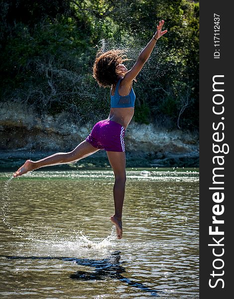 Woman in Blue Sports Bra and Purple Shorts Leaping Above Body of Water