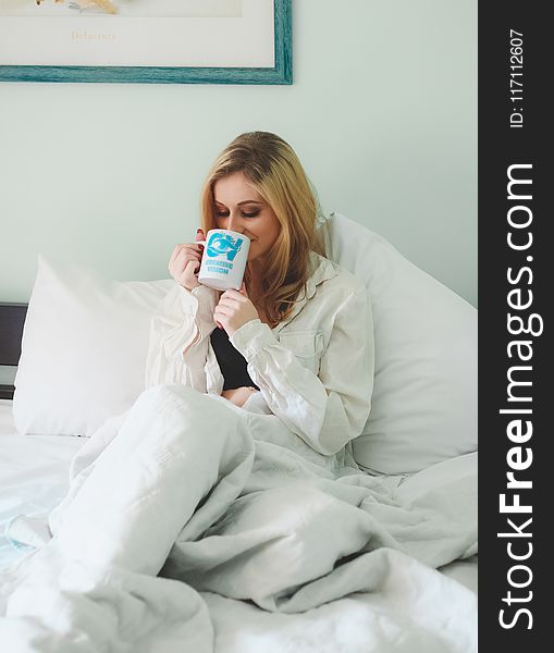 Woman Wearing White Dress Shirt Sitting on Bed While Drinking Through White Ceramic Mug