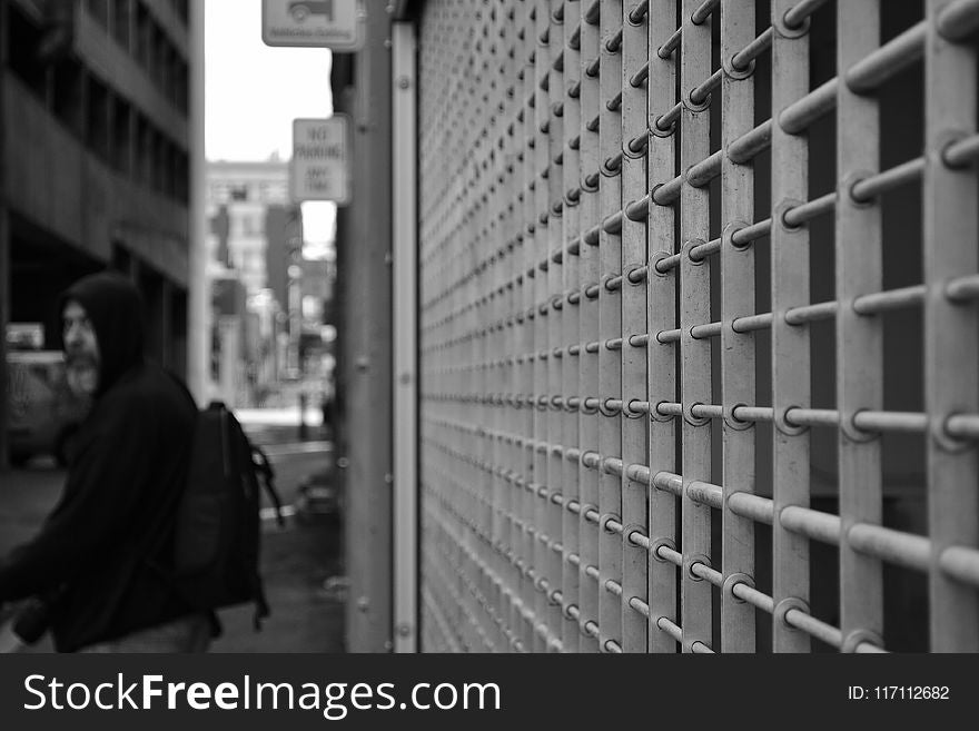 Grayscale Photo of Man in Hoodie in Front of White Window Grill