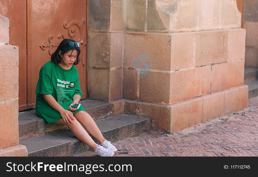Woman Wearing Green Shirt Sitting Near Brown Gate Holding Smartphone