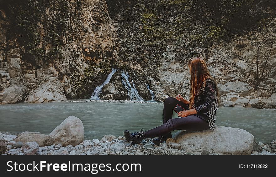 Woman With Brown Hair In Black Jacket Sitting On Rock Near Body Of Water