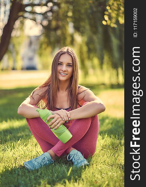 Healthy lifestyle concept. Young girl in sportswear in lotus pose sitting on green grass and holds bottle of water.