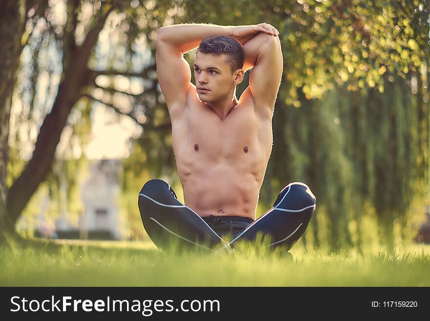 Healthy lifestyle concept. Shirtless young man stretching hands while sitting in lotus pose on a green grass.