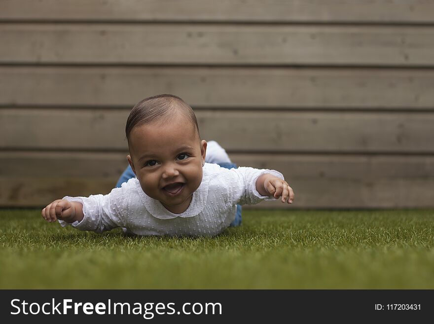 Adorable cute six months old baby girl lying on belly on grass surface with wooden background. Adorable cute six months old baby girl lying on belly on grass surface with wooden background