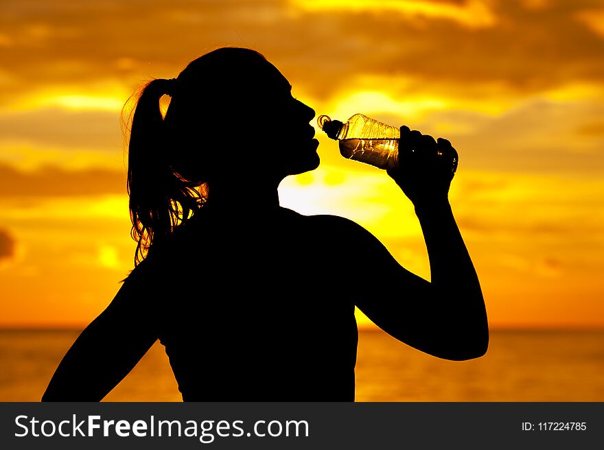 Silhouette of young athletic woman drinking water after outdoors activity in summer sunset time