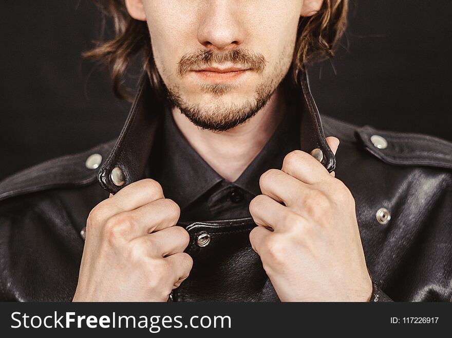 Dramatic fashion model in leather jacket resting elbow on knee while seated on black background. Dramatic fashion model in leather jacket resting elbow on knee while seated on black background