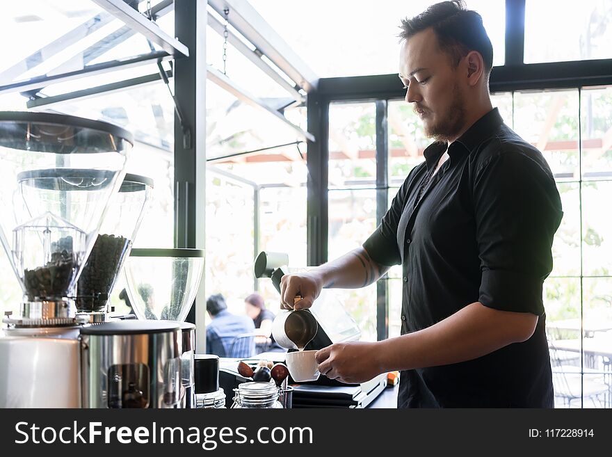 Side view of a young serious bartender pouring fresh milk into a cup of coffee behind the bar counter in a trendy cafeteria